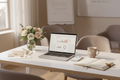 Laptop on a desk with a flower arrangement, coffee cup, and notebook in a bright room.